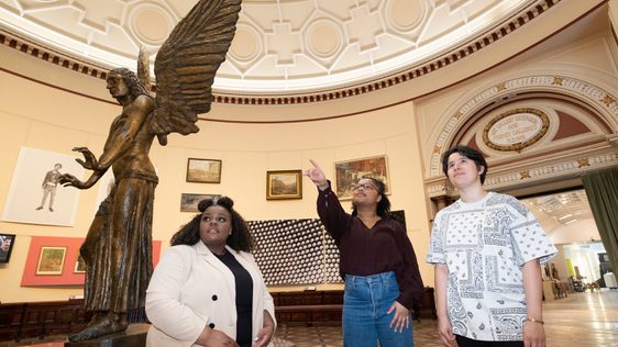 Three people in gallery space, one person is pointing and the others look up in the same direction. A large statue of a winged angel is to the side.