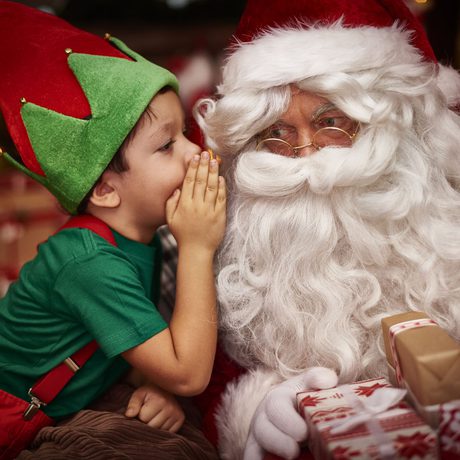 A child dressed as an elf whispering into Santa's ear.