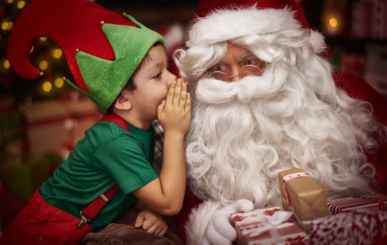 A child dressed as an elf whispering into Santa's ear.