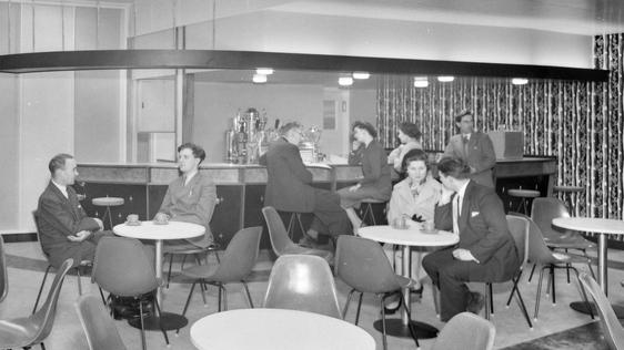 Black and white photo of people sitting small retro looking tea room with plastic chairs and tables.