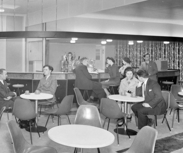 Black and white photo of people sitting small retro looking tea room with plastic chairs and tables.