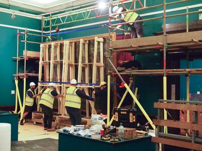 The painting is being moved onto a large wooden frame. There are several men in ghard hats and hi-vis jackets holding the frame.