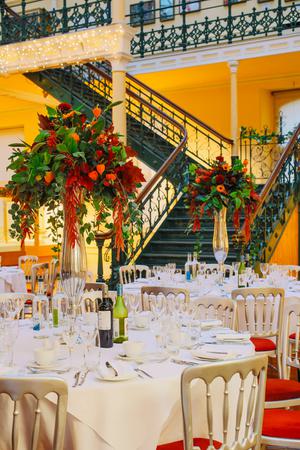 There are several round tables with white tablecloths set up for people to dine. There are gold coloured chairs with red seats. At the centre of the tables are tall glass vases with bouquets of flowers that are red, orange and green.