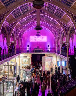 Lots of visitors in two level gallery which has an ironwork ceiling and balcony. The gallery is lit up in pink neon colours.