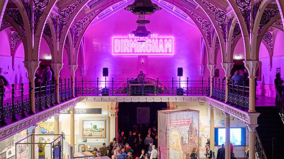 Lots of visitors in two level gallery which has an ironwork ceiling and balcony. The gallery is lit up in pink neon colours.