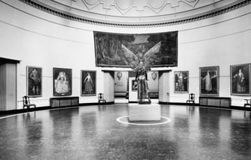 Old black and white photograph showing the statue of Lucifer from behind, with his wings in the air. He is standing in the Round Room gallery on a white plinth. There are historic portraits of people on the walls of the gallery.