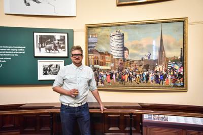 The artist Cold War Steve is standing in a gallery next to his artwork Benny's Babbies, which is hanging on the wall behind him.