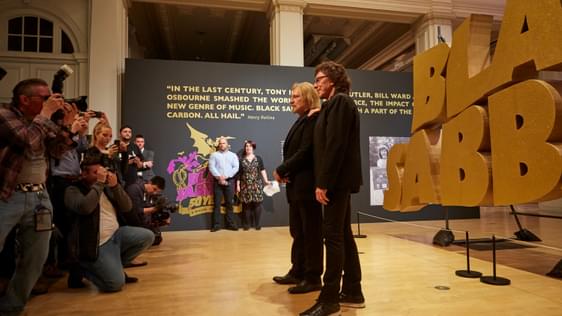 Tony Iommi and Geezer Butler standing in front of large sign that says 'Black Sabbath' in gold letters. They are being photographed by a large group of press photographers.