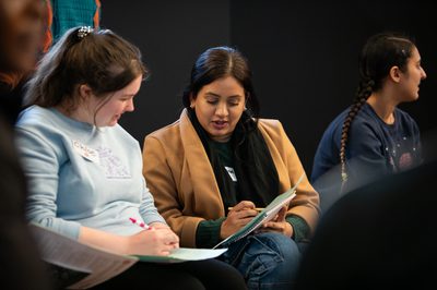 Two people sitting, they are talking and looking at their paper and pens.