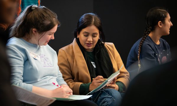 Two people sitting, they are talking and looking at their paper and pens.