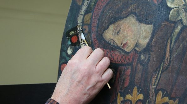 A hand is shown with a brush gently cleaning an artwork. You can see a face of a young man with long hair in the art work.