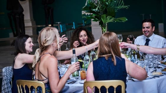 A group of people smiling while sitting at a round table raising their glasses.