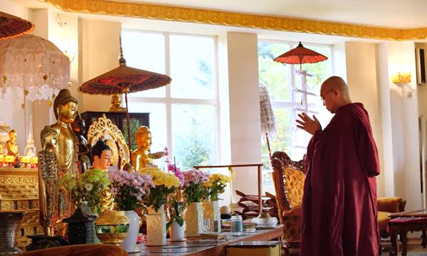 A buddhist monk dressed in red is standing in front of a Buddhist shrine. His hands are closed together in prayer.