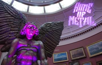 The upper half of the statue of Lucifer is shown lit by a pink light, and on the wall behind there is the projection of the words Home of Metal.