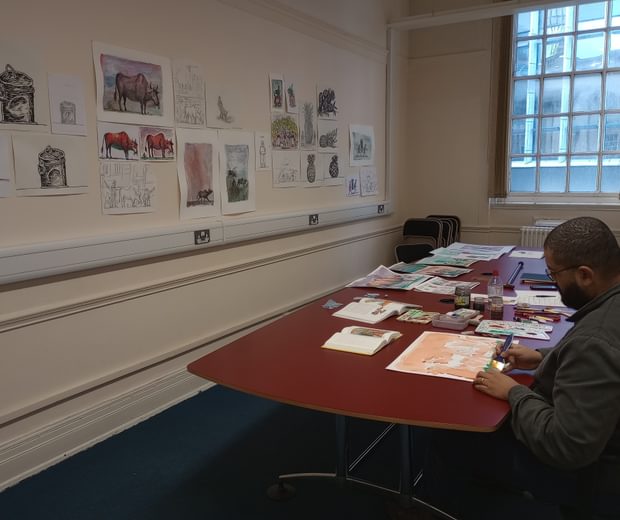 An artist is sitting at a table with a folder of William Blake artworks in front of him.