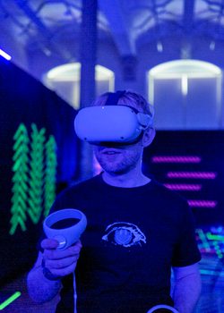 A person wearing a VR headset and controllers in a dimly blue lit room with neon patterns behind them.