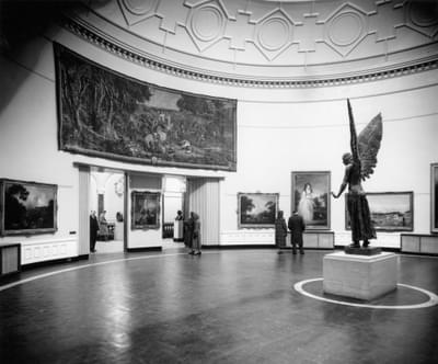 Old black and white photography showing the Round Room gallery. The bronze statue of Lucifer can be seen standing on a white plinth. Lucifer is facing the entrance to the gallery.
