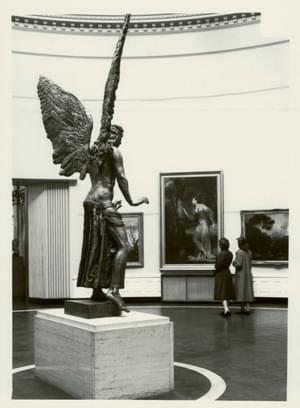 Old black and white photograph showing the state of Lucifer from a side angle, with his wings soaring upwards. In front of him two women are looking at the art on the wall of the gallery.
