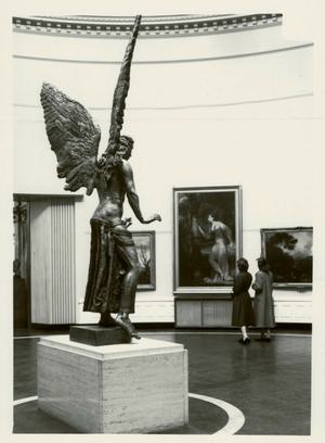 Old black and white photograph showing the state of Lucifer from a side angle, with his wings soaring upwards. In front of him two women are looking at the art on the wall of the gallery.