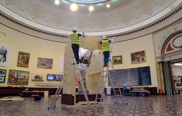 Two men in yellow vests and white hard hats are standing on stepladders and lifting some wood off the box surrounding the statue or Lucifer. Lucifer can be glimpsed in the box.