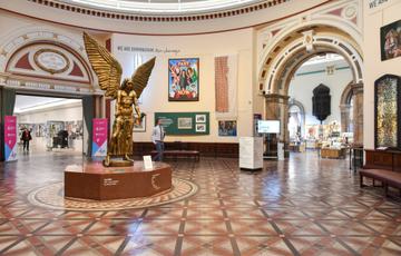 The statue of Lucifer is standing in the middle of the Round Room gallery. The walls are an off white and there are paintings, photographs and textiles hanging from the walls.