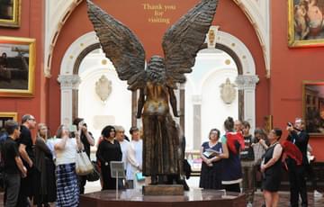 The statue of Lucifer can be seen from the back. In front of him is a group of people listening to a woman talk about the statue. Some of the people are wearing wings.
