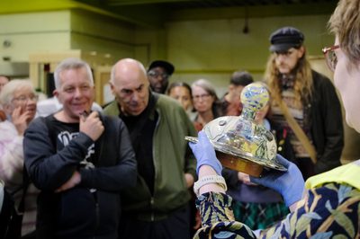 A member of staff holding a object from the collection with visitors looking at it in the background.