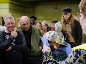 A member of staff holding a object from the collection with visitors looking at it in the background.