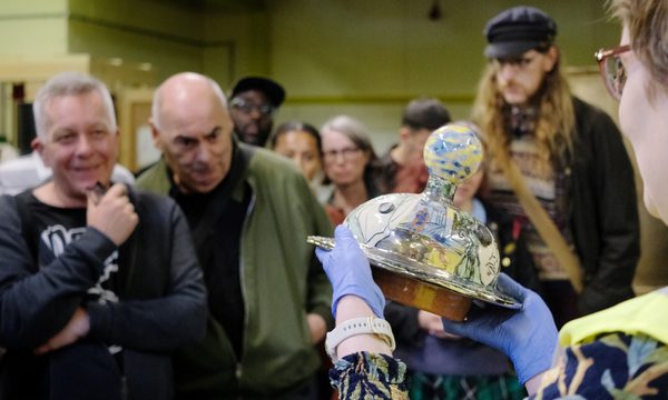 A member of staff holding a object from the collection with visitors looking at it in the background.