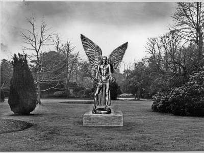 Black and white photograph of a park with grass and trees. An image of the statue of Lucifer has been overlaid over it.