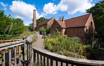Exterior of a brick mill building with chimney, leading to the mill is a footpath with lots of green plants.