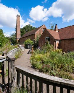 Exterior of a brick mill building with chimney, leading to the mill is a footpath with lots of green plants.