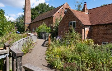 Exterior of a brick mill building with chimney, leading to the mill is a footpath with lots of green plants.