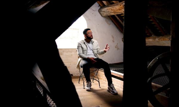 Poet Casey Bailey is sitting on a chair at Sarehole Mill. He is viewed through the dark wood and shadows from inside the Mill, making him look as if he is in a frame. Behind him is a white wall and light coming in through a window.