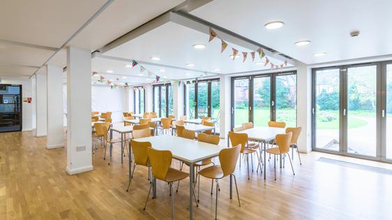 Tables and chairs set up in a room. The wall is made up of glass doors annd you can see the grass outside through them.
