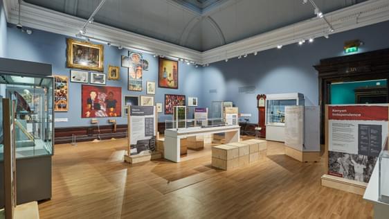 A room in a gallery filled with art, a variety of paintings are on the wall and display cases of objects around the edge.