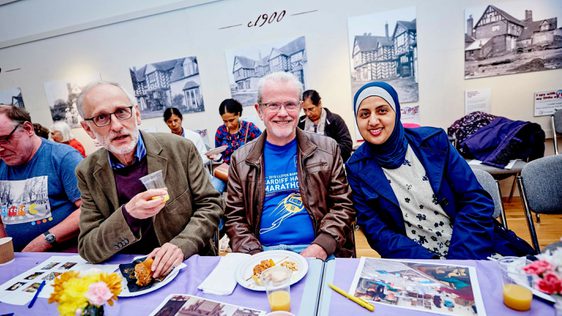 Two men and one female sitting down at a table with food and activities on the table, one man is holding a drink