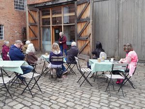 A group of people seated outside listening to a person giving who is standing a talk.