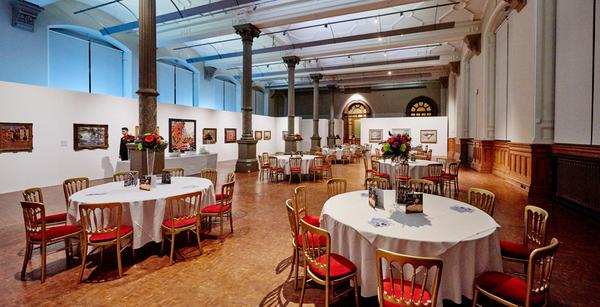 Large room set up for an event. There are several round tables with white tablecloths and gold coloured chairs with red seating. There are multi-coloured flowers in tall vases in the centre of the tables.
