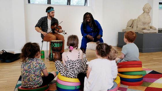 Two musicians with violin and drum perform a piece of music to a group of children sitting on the floor.