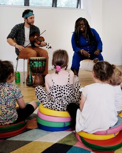 Two musicians with violin and drum perform a piece of music to a group of children sitting on the floor.