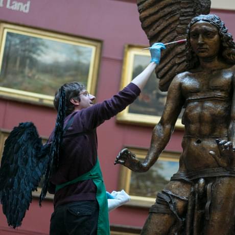 A man wearing black wings is cleaning the statue of Lucifer's face with a small brush.