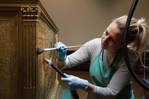 A conservator holding brush and vacuum cleaner attachment, gently dusting a piece of furniture.