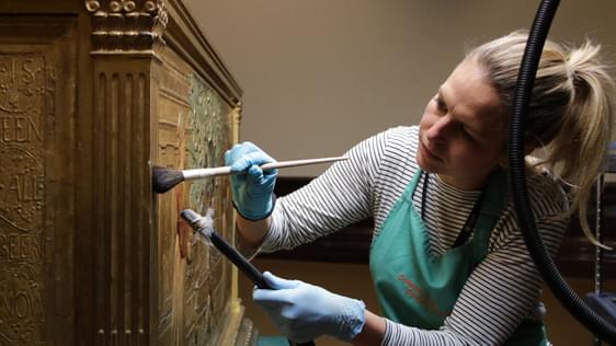 A conservator holding brush and vacuum cleaner attachment, gently dusting a piece of furniture.