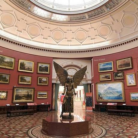 The statue of Lucifer is in the cente of the Round Room gallery. The red walls of the gallery are covered in paintings that are hung three or four deep. The round glass skylight that hangs above the gallery can be partially seen.