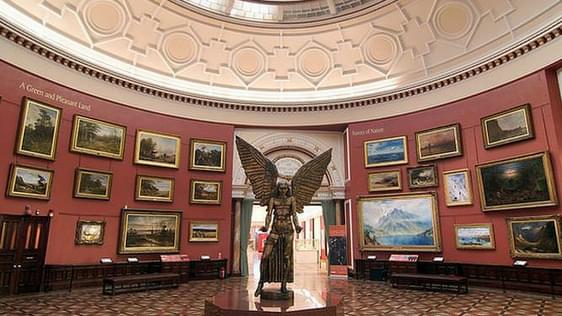 The statue of Lucifer is in the cente of the Round Room gallery. The red walls of the gallery are covered in paintings that are hung three or four deep. The round glass skylight that hangs above the gallery can be partially seen.