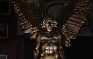 Close up of Lucifer showing his face, upper body and wings. He is partially lit and the rest of the gallery is dark.