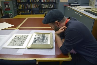 An artist is sitting at a table with a folder of William Blake artworks in front of him.
