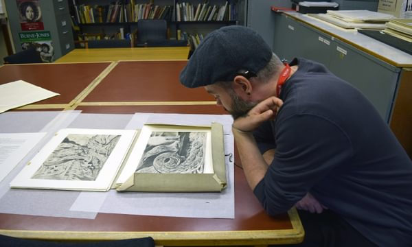 An artist is sitting at a table with a folder of William Blake artworks in front of him.