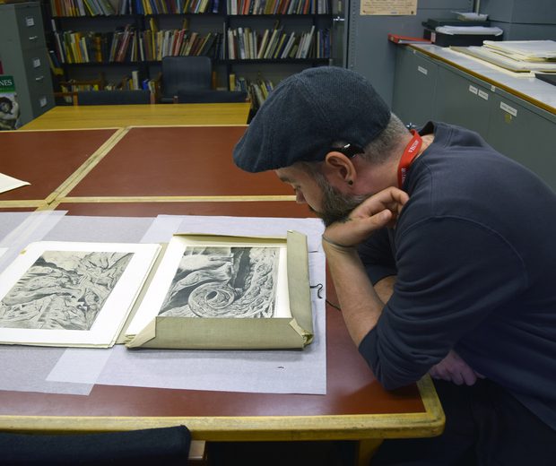 An artist is sitting at a table with a folder of William Blake artworks in front of him.